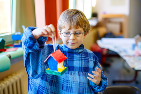 Little school kid boy with glasses coloring bird house with watercolors in labor classroom. child...
