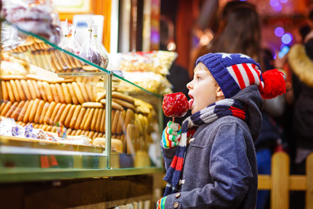 Little cute kid boy eating sugar apple near sweet stand with gingerbread and nuts. Happy child on...