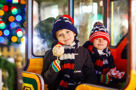 Two little kids boys on a carousel at Christmas funfair or market, outdoors. Happy children having fun and eating sweets. Traditional xmas market in Germany, Europe. Holiday, lifestyle concept.の写真素材