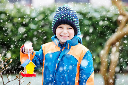 Little kid boy feeding birds in winter. Cute happy preschool child hanging colorful selfmade bird house on tree on frosty cold day. Preschooler in colorful wam clothes. Nature, empathy with animals.の写真素材