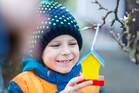 Little kid boy feeding birds in winter. Child hanging colorful selfmade bird house on tree on frosty...
