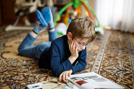 Little blond kid boy with eyeglasses reading book on floor at home