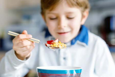 little blond school kid boy eating cereals with milk and berries, fresh strawberry for breakfast