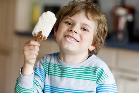 Little blond kid boy with curly hairs eating ice cream popsicle with chocolate at homeの写真素材