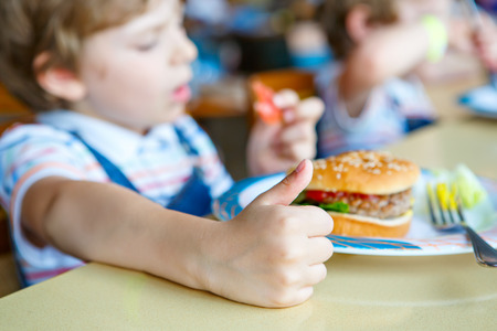 Cute healthy preschool boy eats hamburger sitting in school canteenの写真素材