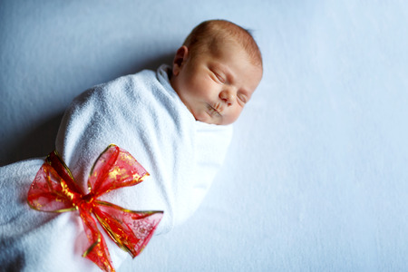 One week old newborn baby sleeping wrapped in white blanket with red bowの写真素材