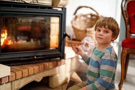 Little cute kid boy sitting by fireplace at home and helping with watching out for fireの写真素材