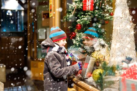 Funny happy child in fashion winter clothes making window shopping decorated with gifts, xmas treeの写真素材
