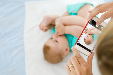 Parent taking photo of a baby with smartphone. Adorable newborn child taking foot in mouthの写真素材