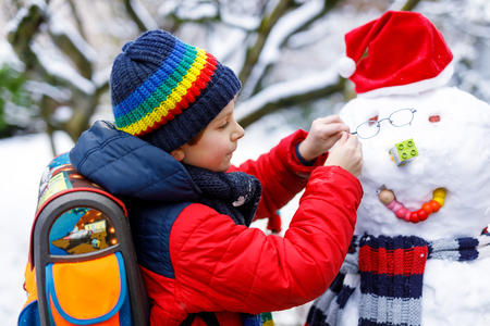 Little school kid boy in colorful clothes, with glasses and backpack having fun with snowman after elementary school endの写真素材
