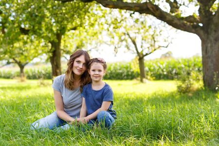 Beautiful young mother and little daughter sitting on green grass and restingの写真素材