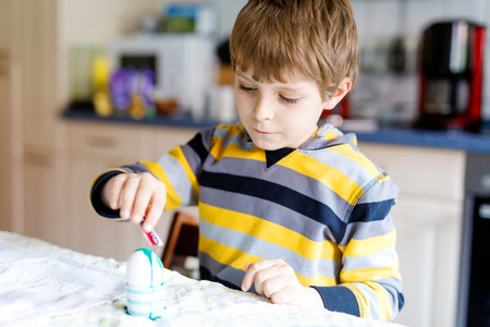 Little blond kid boy coloring eggs for Easter holiday in domestic kitchen, indoors.の写真素材