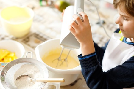 Beautiful funny blond little kid boy baking chocolate cake and tasting dough in domestic kitchenの写真素材