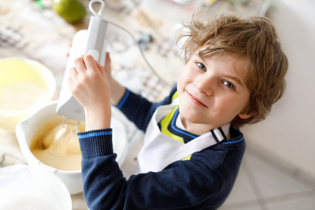 Beautiful funny blond little kid boy baking chocolate cake and tasting dough in domestic kitchenの写真素材