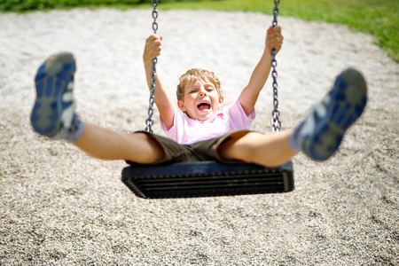 Funny kid boy having fun with chain swing on outdoor playground while being wet splashed with waterの写真素材