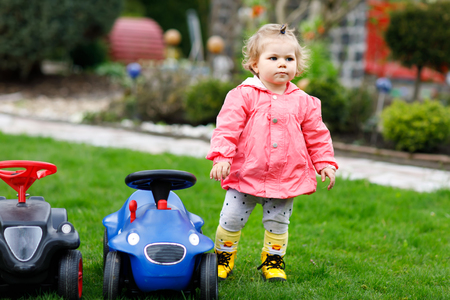Cute little baby girl playing with two toy cars in garden. Adorable toddler child having fun. Girl in colorful fashion clothes.の写真素材