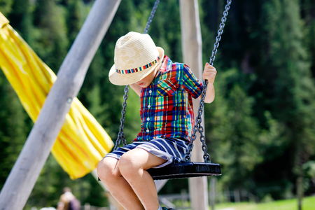 Funny kid boy having fun with chain swing on outdoor playground while being wet splashed with waterの写真素材