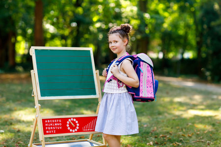Happy little kid girl standing by desk with backpack or satchel. Schoolkid on first day of...