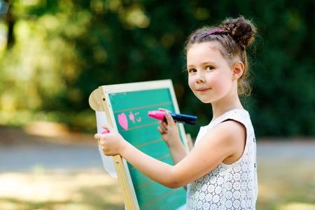 Happy little kid girl standing by big chalk desk Preschool or schoolkid on first day of elementary class. Back to school concept. Healthy child writing and painting on desk outdoors. Copyspace on deskの写真素材