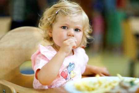 Adorable toddler girl eating healthy vegetables and unhealthy french fries potatoes. Cute happy baby child taking food from dish at daycare or nursery canteen.の写真素材