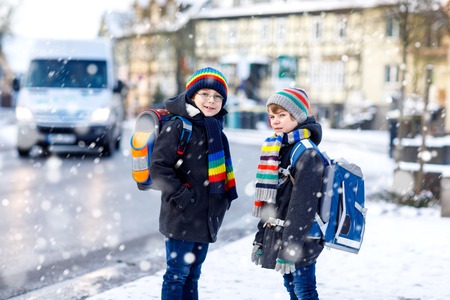 Two little kids boys of elementary class walking to school during snowfall. Happy children having fun and playing with first snow. Siblings ans friends with backpack in colorful winter clothes.の写真素材