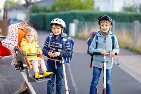 Two little school kid boys and cute toddler girl going to school. Tiny child sister sitting in pram. Brothers riding on scooter. Happy healthy amily of three children.の写真素材
