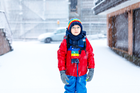 Little school kid boy of elementary class walking to school during snowfall. Happy child having fun and playing with first snow. Student with eye glasses backpack in colorful winter clothesの写真素材