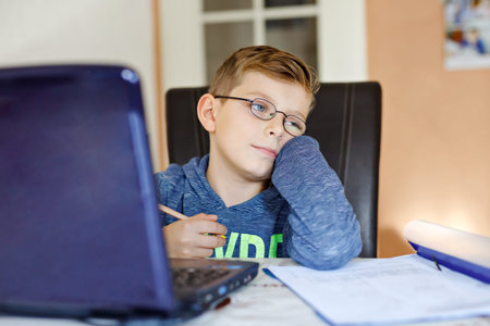 Tired sad kid boy with glasses making school homework at home with notebook. Depressed child writing...