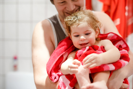 Father holding cute little toddler girl wrapped in towel after taking bath. Happy healthy baby child with wet hairs looking in mirror in bathroom and laughing. Family relationship of daughter and dadの写真素材