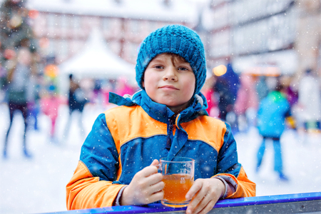 Happy little kid boy in colorful warm clothes on skating rink of Christmas market or fair drinking hot punch or chocolate. Healthy child having fun on ice skate. people having active winter leisureの写真素材