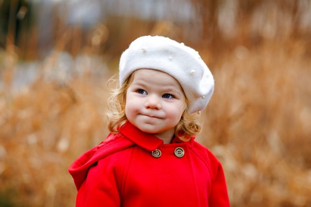 Outdoor portrait of little cute toddler girl in red coat and white fashion hat barret. Healthy happy baby child walking in the park on cold day. Fashion stylish clothes for kids.の写真素材