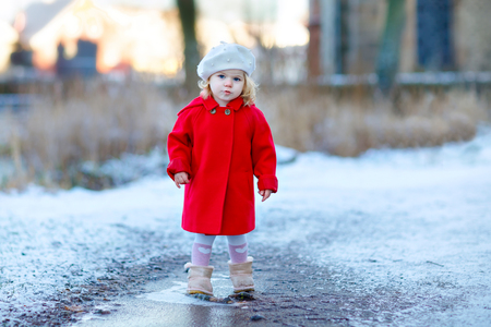 Outdoor winter portrait of little cute toddler girl in red coat and white fashion hat barret. Healthy happy baby child walking in the park on cold day with snow and snowfall. Stylish clothes for kids.の写真素材
