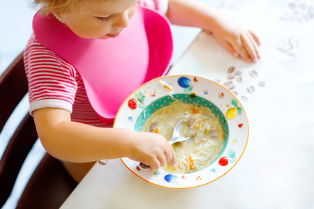 Close-up baby girl eating from spoon vegetable noodle soup. food, child, feeding and development concept. closeup of toddler, daughter with spoon sitting in highchair and learning to eat by itselfの写真素材