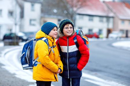 Two little kids boys of elementary class walking to school during snowfall. Happy children having fun and playing with first snow. Siblings and best friends with backpack in colorful winter clothes.の写真素材