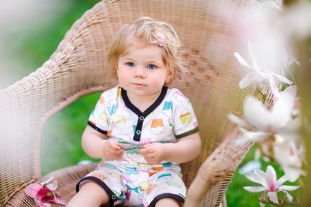 Cute little baby girl sitting on big chair in garden. Beautiful happy smiling toddler with blooming pink magnolia tree on background. Healthy child enjoying spring season.の写真素材