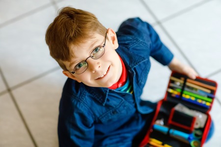 Happy little school kid boy searching for a pen in pencil case. Healthy schoolchild with glasses grab thinks for lessons in elementary class.の写真素材