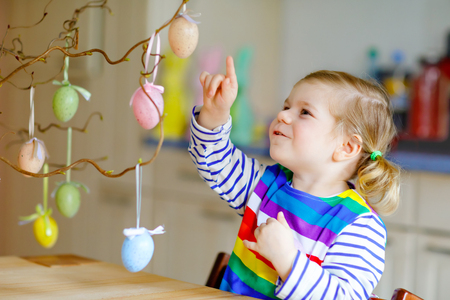 Cute little toddler girl decorating tree bough with colored pastel plastic eggs. Happy baby child having fun with Easter decorations. Adorable healthy smiling kid in enjoying family holidayの写真素材