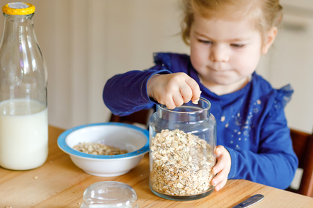 Adorable toddler girl eating healthy oatmeals with milk for breakfast. Cute happy baby child in colorful clothes sitting in kitchen and having fun with preparing oats, cereals. Indoors at homeの写真素材