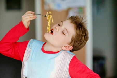 Cute healthy preschool kid boy eats pasta noodles sitting in school or nursery cafe. Happy child eating healthy organic and vegan food in restaurant or at home. Childhood, health conceptの写真素材