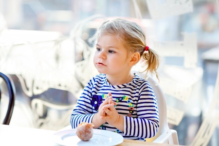 Cute little adorable toddler girl sitting in indoor cafe restaurant. Happy healthy baby child eating bread or sweet scone on sunny day.の写真素材