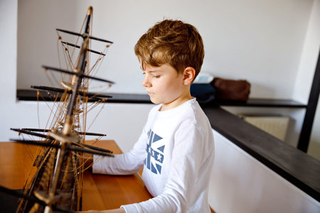 Little blond school kid boy playing with sailing ship model indoors. Excited child with yacht having fun after school at home. Happy boy building and creating toysの写真素材