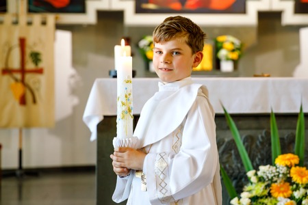 Little kid boy receiving his first holy communion. Happy child holding Christening candle. Tradition in catholic curch. Kid in a white traditional gown in a church near altar.の写真素材
