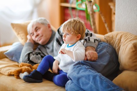 cute little toddler boy and grandfather watching together tv show. Baby grandson and happy retired senior man sitting together at home with cartoons on television. Calm family time.の写真素材