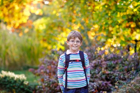 Happy little kid boy with glasses and backpack or satchel on his first day to school on sunny autumn day. Child outdoors with yellow and read maple trees on background, Back to school conceptの写真素材