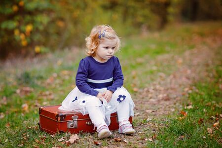 Cute little toddler girl sitting on suitcase in autumn park. Happy healthy baby enjoying walking with parents. Sunny warm fall day with child. Active leisure and activity with kids in nature.の写真素材