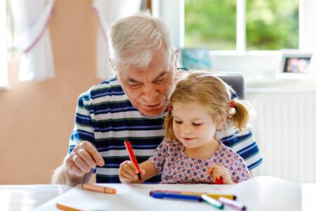 Cute little baby toddler girl and handsome senior grandfather painting with colorful pencils at home. Grandchild and man having fun togetherの写真素材