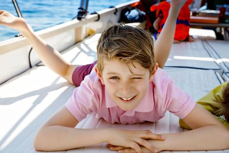 Happy blonde kid boy enjoying sailing boat trip. Family vacations on ocean or sea on sunny day. Healthy beautiful school child smiling and having fun on yacht. Coastline with villages and natureの写真素材