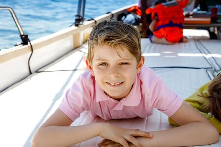 Happy blonde kid boy enjoying sailing boat trip. Family vacations on ocean or sea on sunny day. Healthy beautiful school child smiling and having fun on yacht. Coastline with villages and natureの写真素材