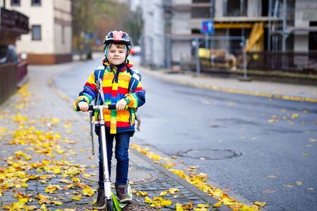 cute little school kid boy riding on scooter on way to elementary school. Child with safety helmet, school bag on rainy autumn day. Traffic in the city and schoolchildren.の写真素材