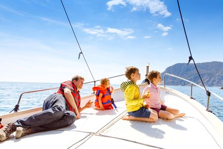 Two little kid boys, father and toddler girl enjoying sailing boat trip. Family vacations on ocean or sea on sunny day. Children smiling. Brothers and sister, siblings and dad having fun on yacht.の写真素材